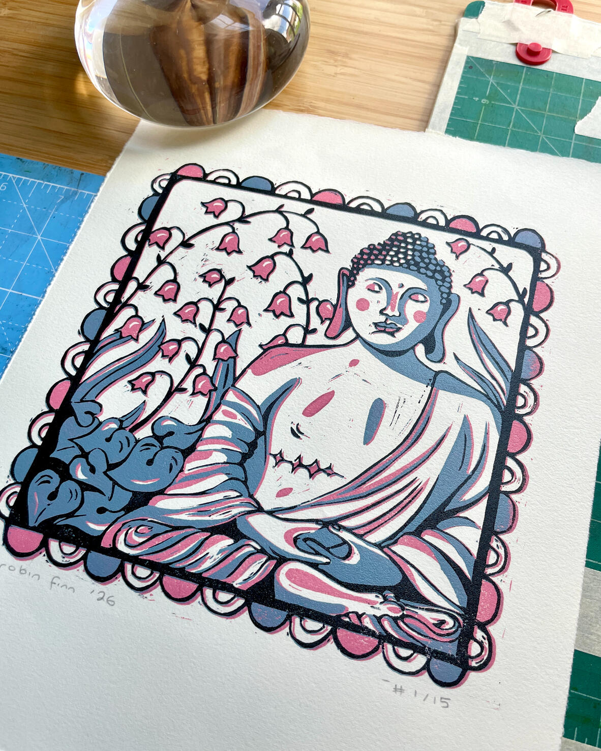 Trans Buddha | buy prints Linocut print of a Buddha statue with top surgery scars, in a garden of Lily of the Valley, surrounded by a decorative border. Printed in pink, blue, and black. Pictured with printmaking tools.