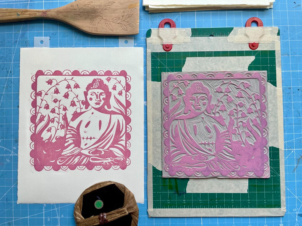 Pink Layer A freshly pulled, unfinished linocut print of a Buddha statue with top surgery scars, next to the linoleum block on a cutting mat marked with tape and registration tabs to line up the paper. There are also two tools on the table: a bamboo baren and a woode