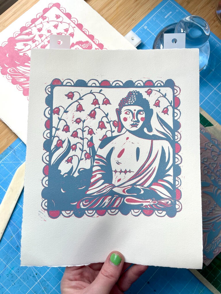 Blue Layer Unfinished, two color linocut block print of a buddha statue with top surgery scars in a garden of lily of the valley. Printmaking tools in the background.