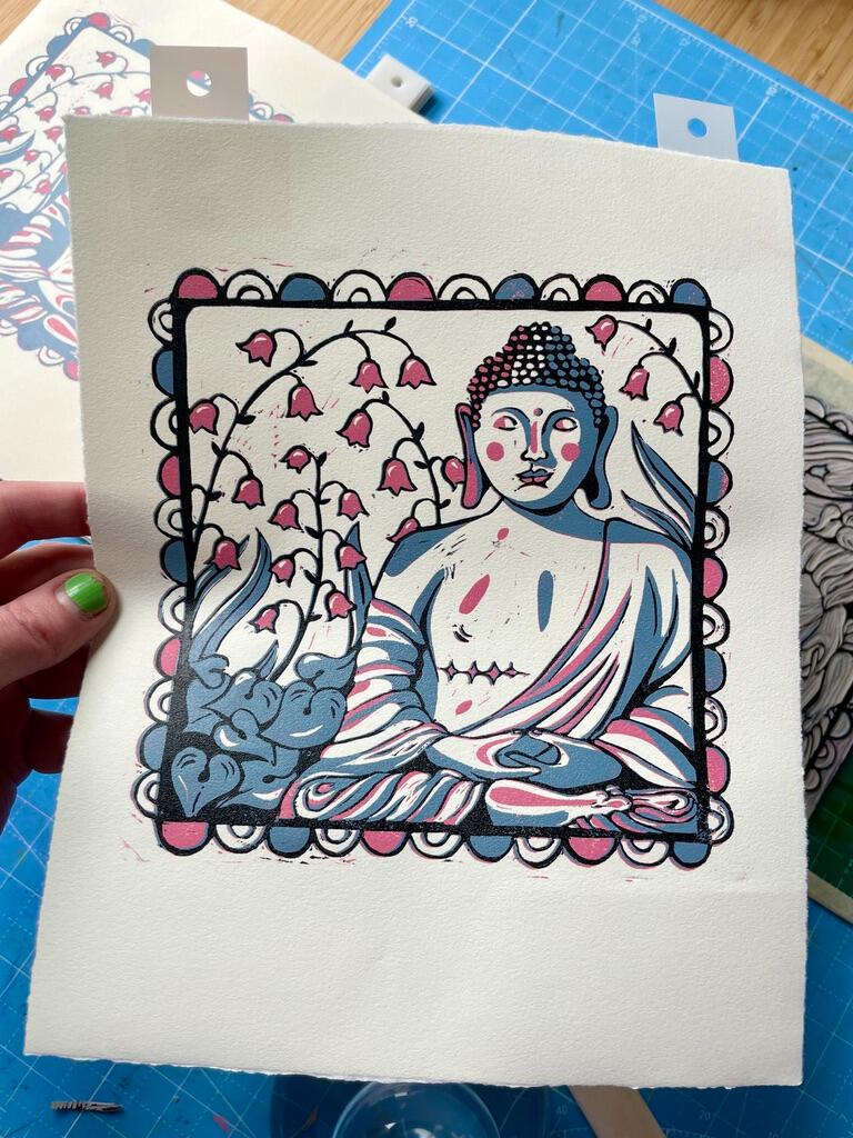 Final Layer A linocut print of a Buddha statue with top surgery scars in a garden of lily of the valley, with a decorative border. Printmaking supplies in the background.