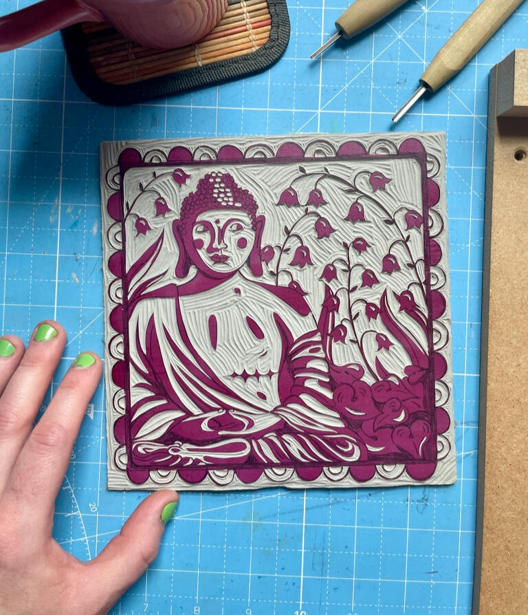 Block A carved linoleum block on a cutting mat next to carving tools, a mug of tea, and Robin's hand. The design is a buddha statue with top surgery scars surrounded by lily of the valley flowers, leaves, and a decorative border.