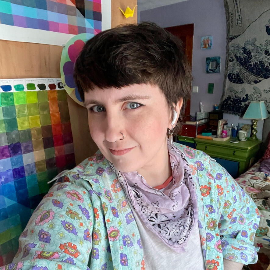 Robin, a person with light skin and short dark brown hair, smiles at the camera from their studio bedroom. They are wearing an oversize paisley shirt, a bandana necktie, and winged eyeliner.